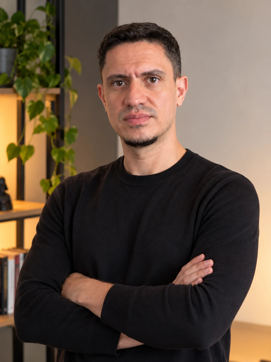 Portrait of Leonardo Carvalho with arms crossed, in front of a shelf with plants.